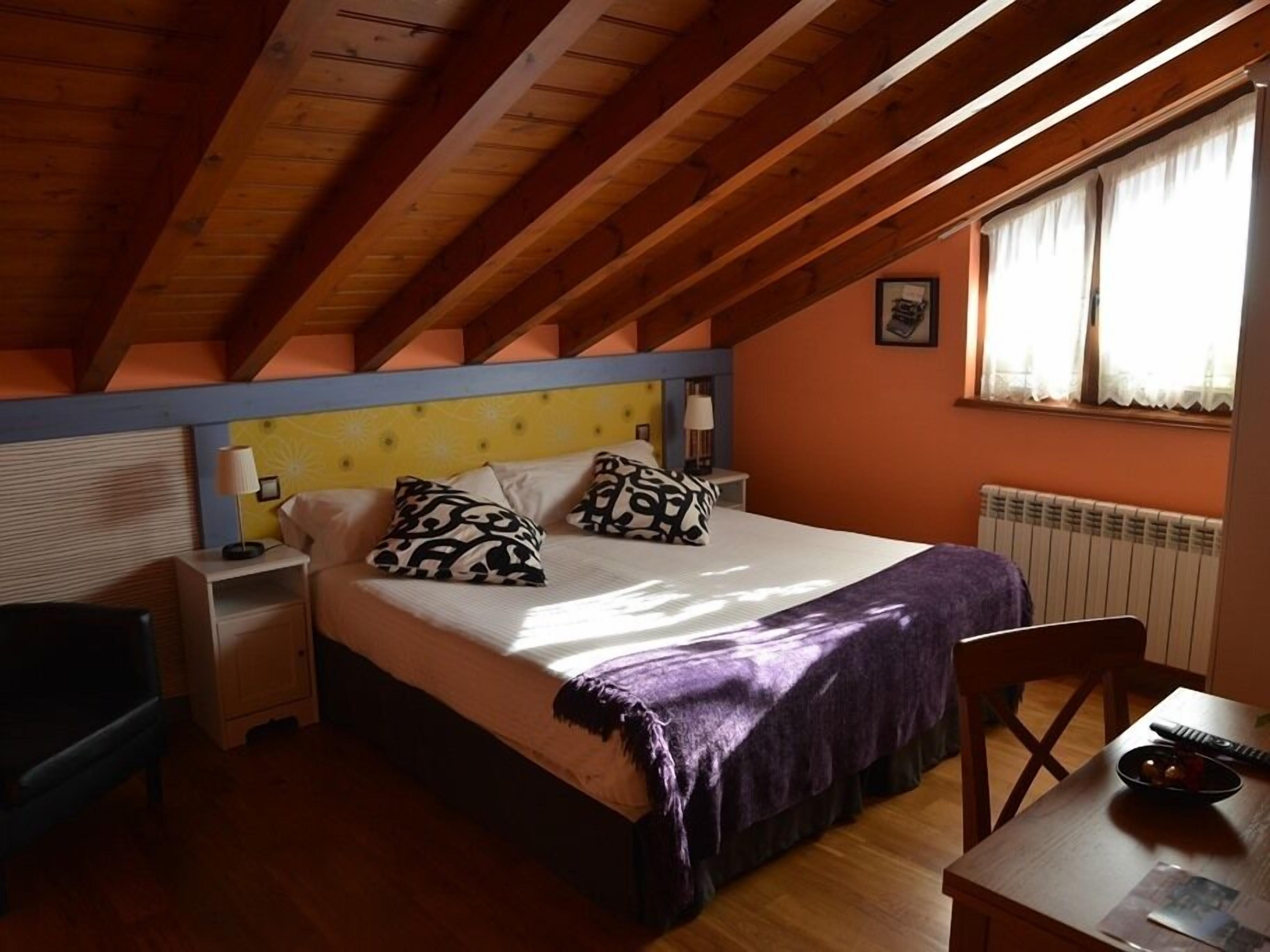 A bedroom with a sloped ceiling, a bed with patterned pillows, a bedside table, a chair, and a table with a bowl and remote control.