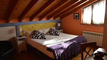 A bedroom with a sloped ceiling, a bed with patterned pillows, a bedside table, a chair, and a table with a bowl and remote control.