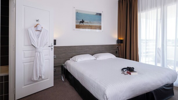 A hotel room with a bed, a robe hanging on the door, a painting of a beach scene, and a window with curtains. - Golden Tulip La Baule Hôtel & Résidence (La Baule-Escoublac)