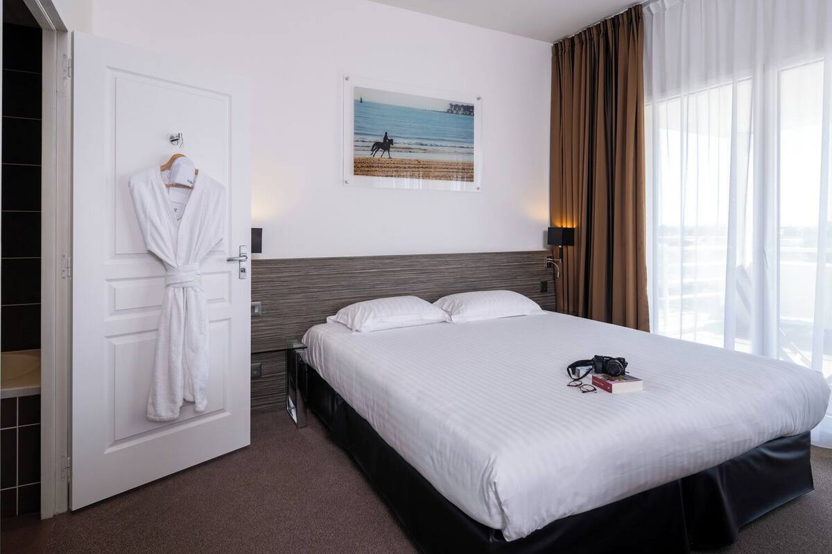 A hotel room with a bed, a robe hanging on the door, a painting of a beach scene, and a window with curtains.
