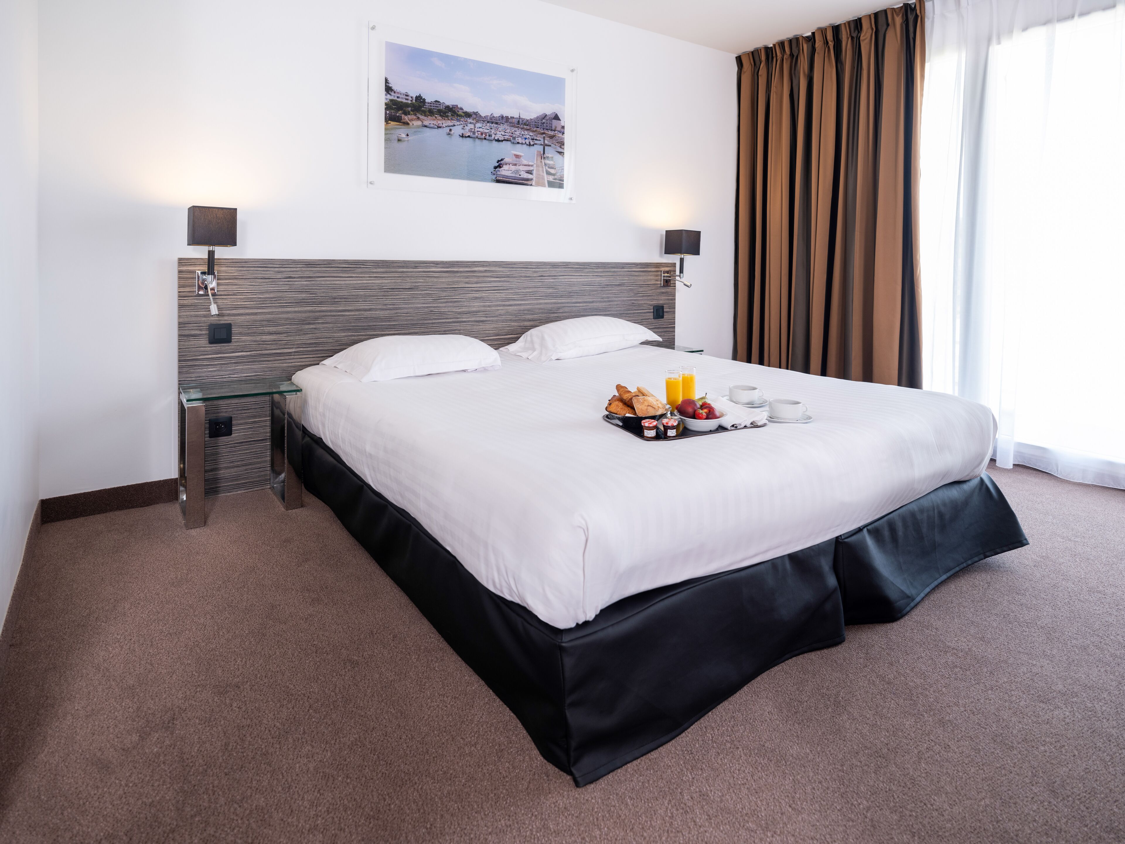 Hotelkamer met een groot bed, nachtkastje, een ingelijst schilderij aan de muur en een raam met gordijnen.