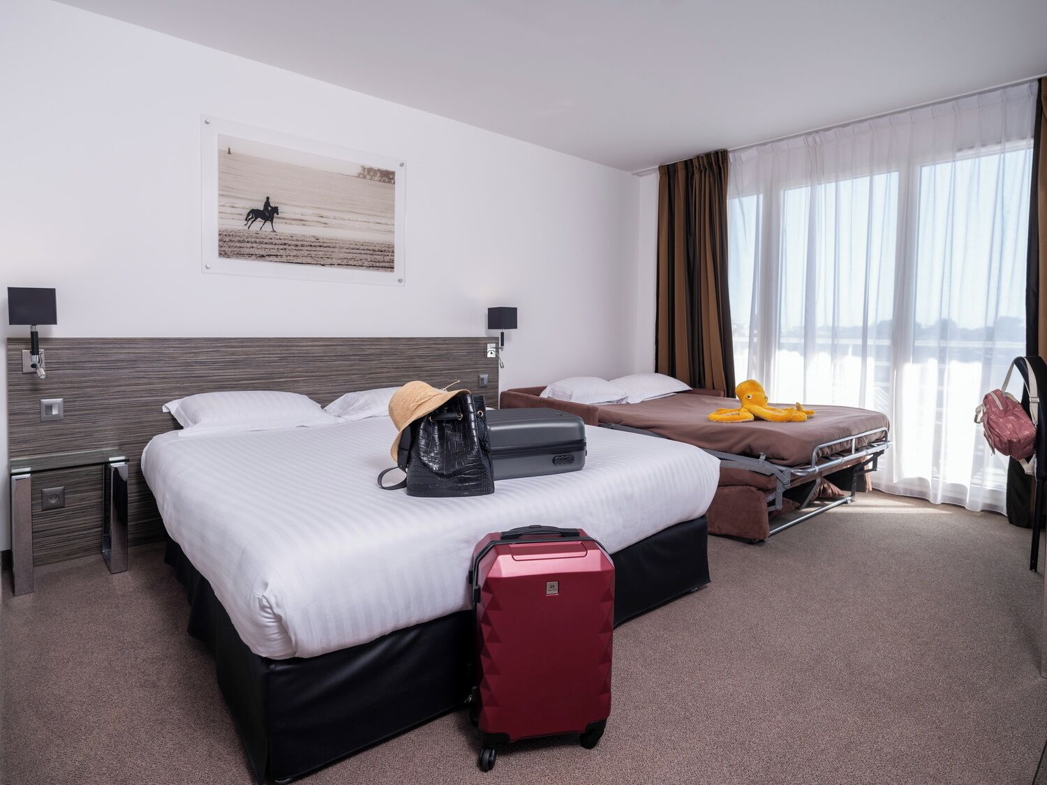 Quarto de hotel com duas camas, uma mala vermelha, uma bolsa preta e um brinquedo de polvo amarelo sobre uma das camas.