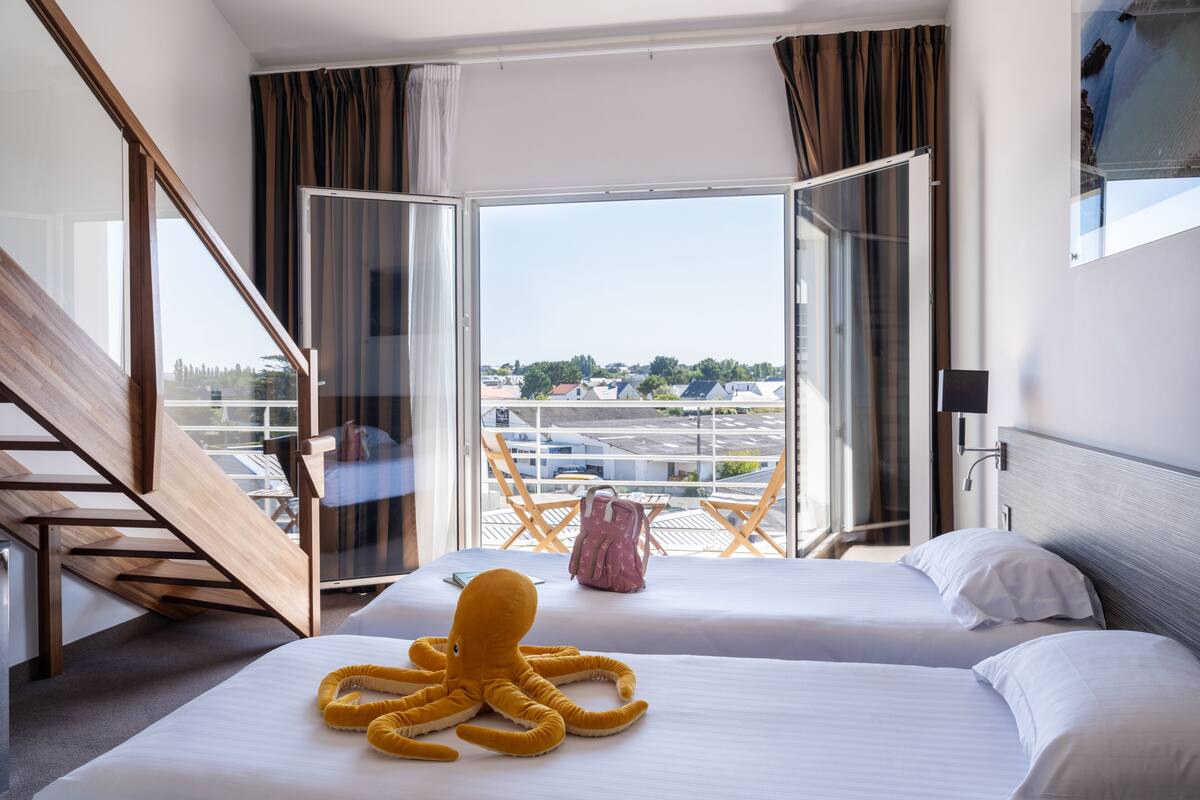 A hotel room with two beds, a large window with curtains, a view of the city, and a wooden staircase.