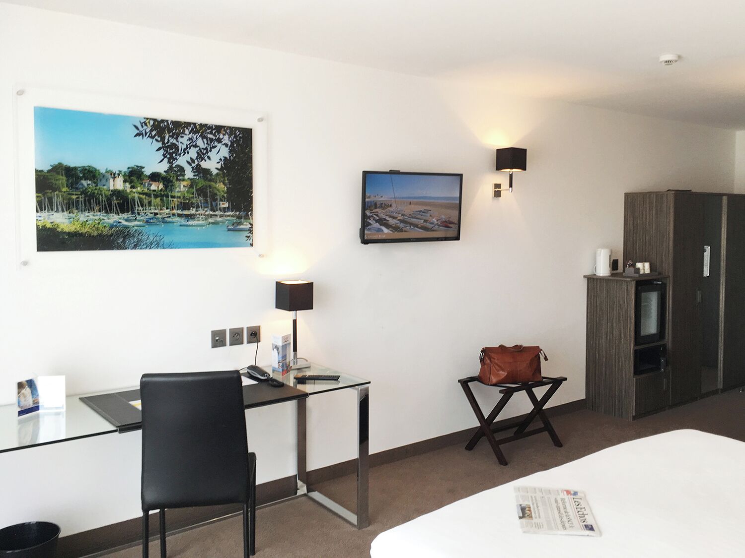 A hotel room with a bed, a desk with a chair, a small table with a bag, a TV, and two wall-mounted lamps.