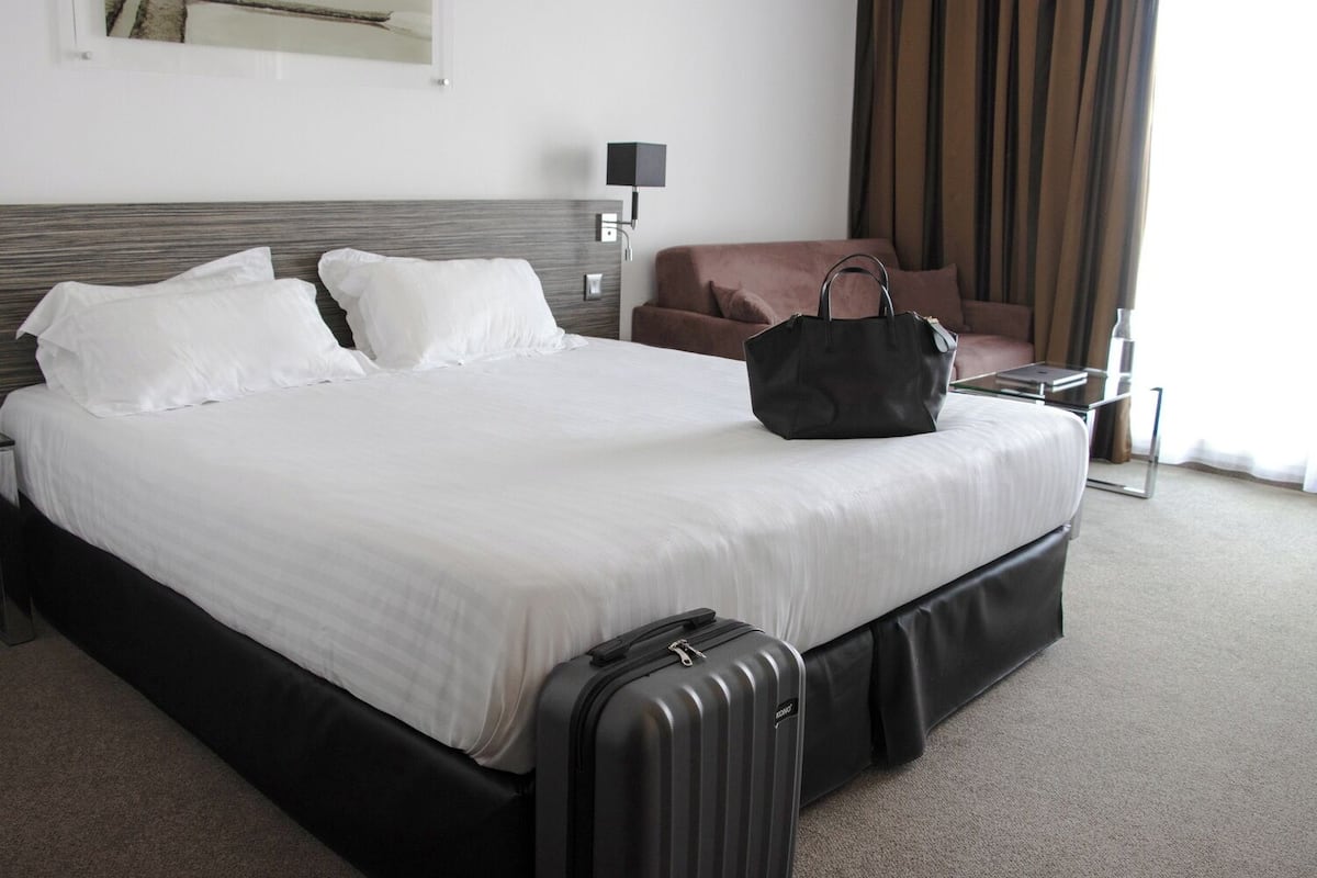 A hotel room with a bed, a suitcase, a black handbag, a brown armchair, a lamp, and a window with curtains.