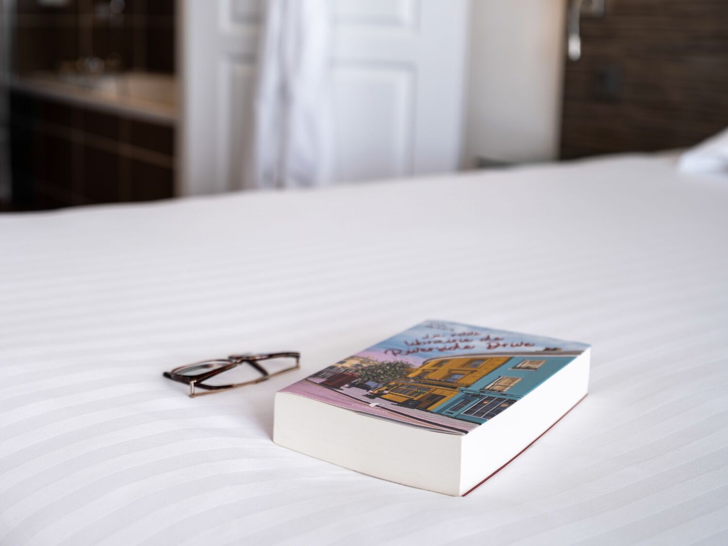 Un livre et des lunettes sur un lit recouvert d’une couverture blanche.