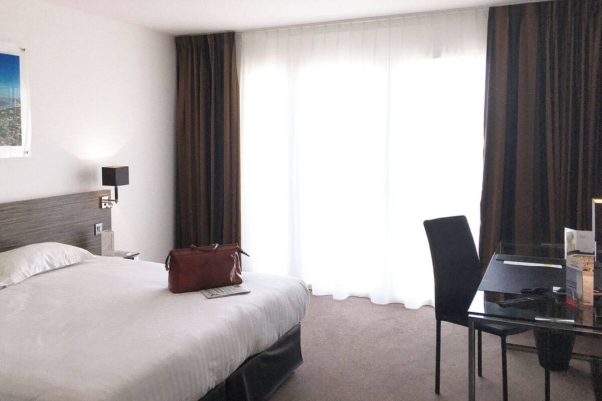 A hotel room with a bed, a desk with a chair, a lamp, and a window with curtains.