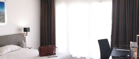 A hotel room with a bed, a desk with a chair, a lamp, and a window with curtains.