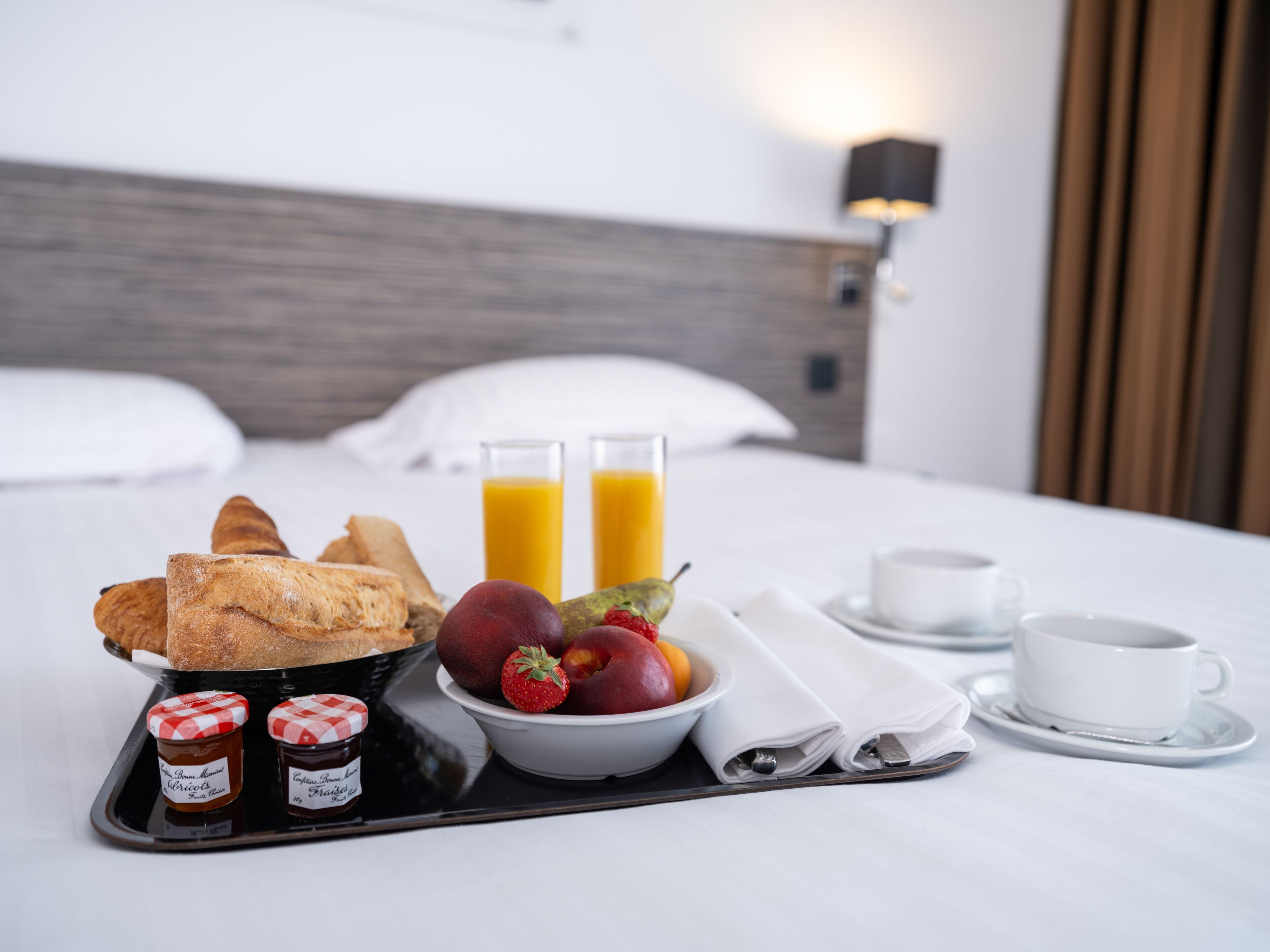 Ein ordentlich gedecktes Frühstücksbrett mit Croissants, Früchten und Marmelade auf einem Bett mit weißen Bettwäsche und einem hölzernen Kopfteil.