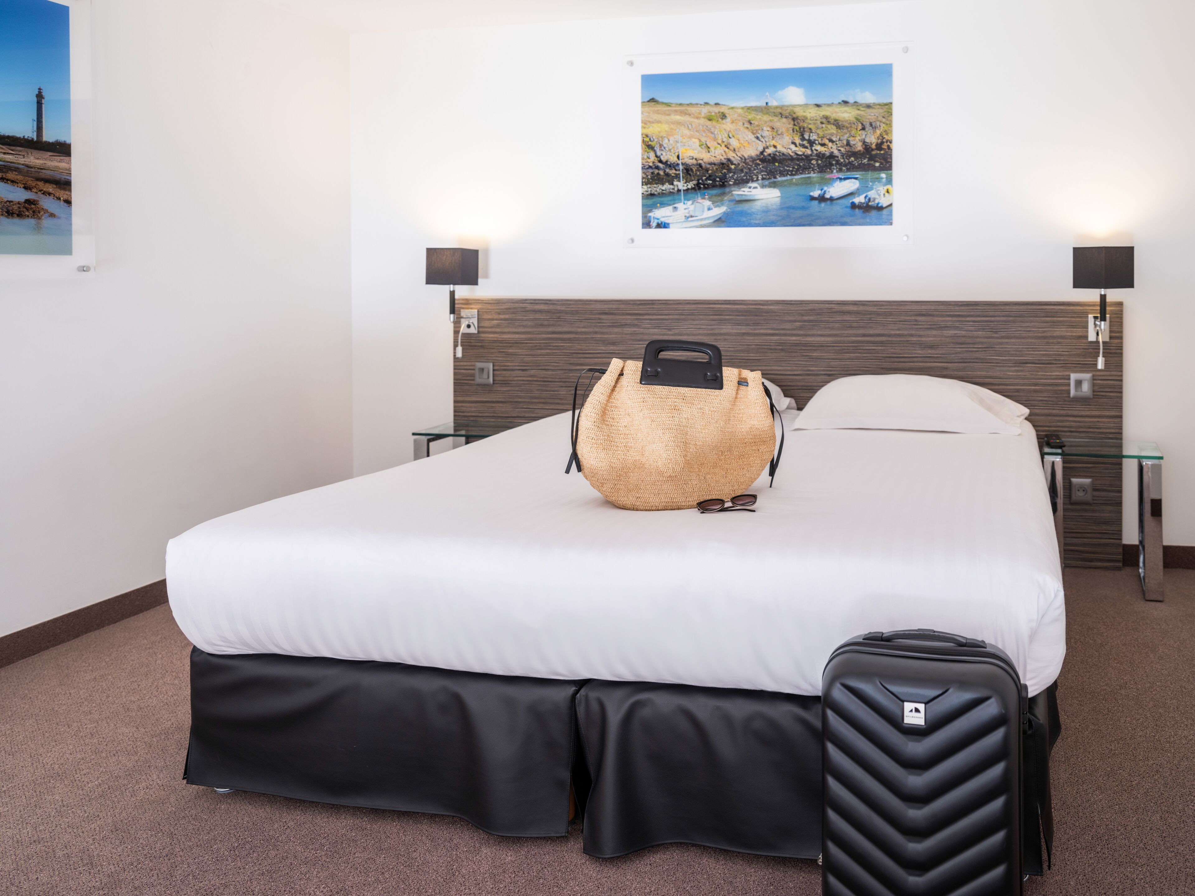 A hotel room with a bed, a suitcase, a handbag, and a painting of a coastal scene.