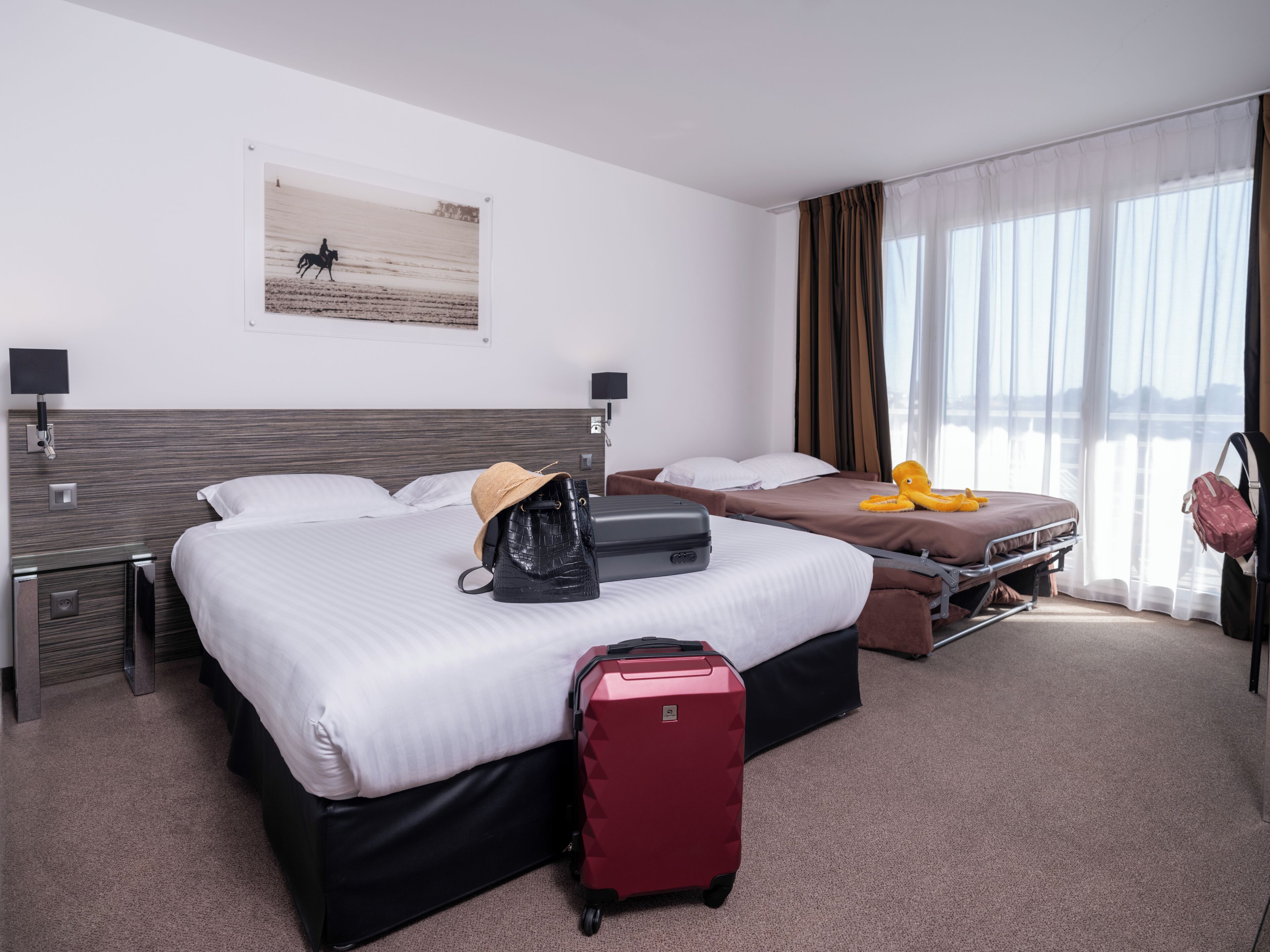Habitación de hotel con dos camas, una maleta roja, un bolso negro y un peluche amarillo sobre una de las camas.