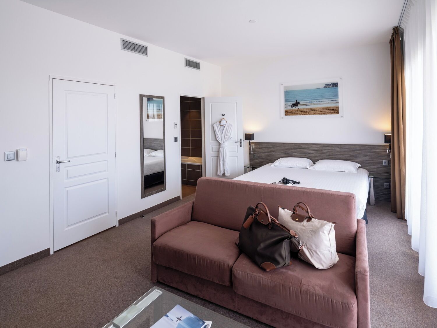 A hotel room with a bed, a sofa, a mirror, a robe, and a view of the sea.