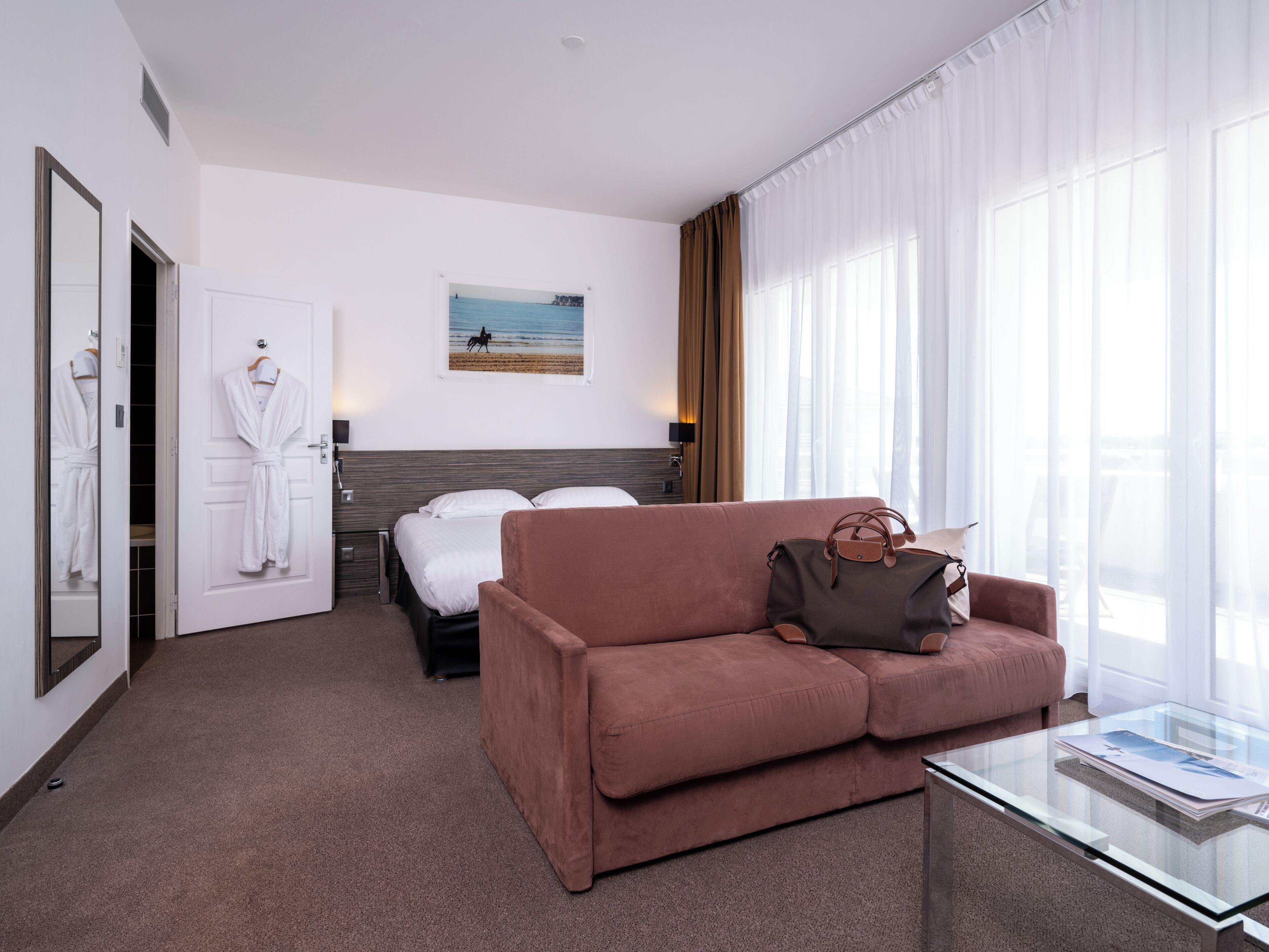 Una habitación de hotel con una cama, un sofá, una mesa de centro de vidrio, un cuadro en la pared y una ventana con cortinas.