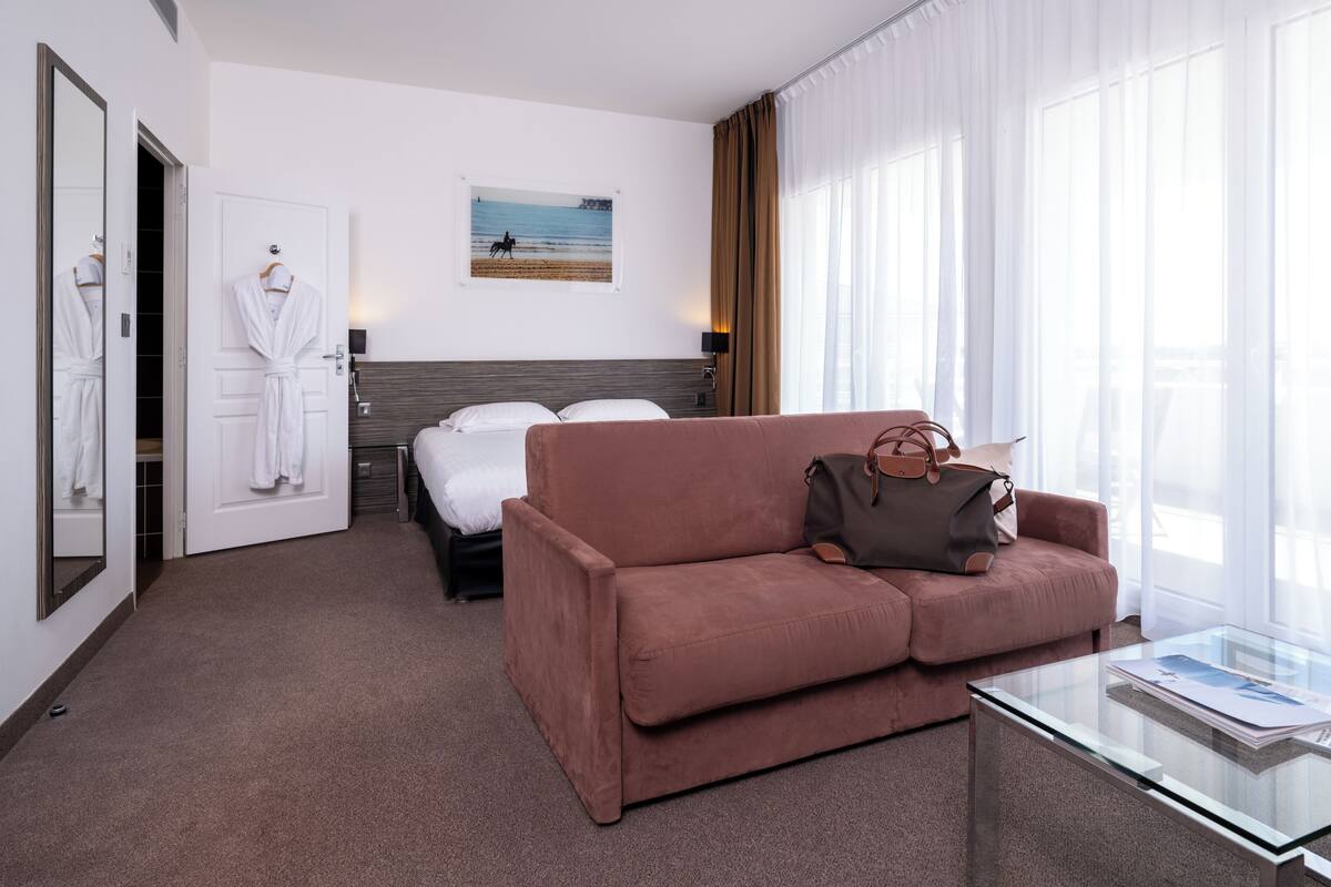 A hotel room with a bed, a sofa, a glass coffee table, a painting on the wall, and a window with curtains.