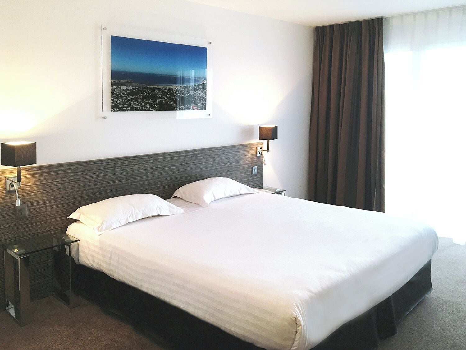 Una habitación de hotel con una cama grande, dos mesitas de noche, una lámpara de pared y un cuadro enmarcado sobre la cama.