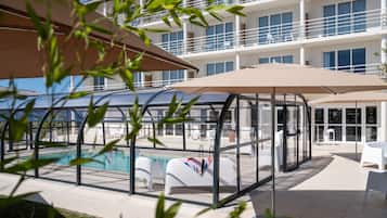 A modern hotel with a glass-enclosed pool area, outdoor seating, and a multi-story building with balconies.