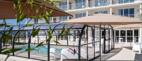 A modern hotel with a glass-enclosed pool area, outdoor seating, and a multi-story building with balconies.
