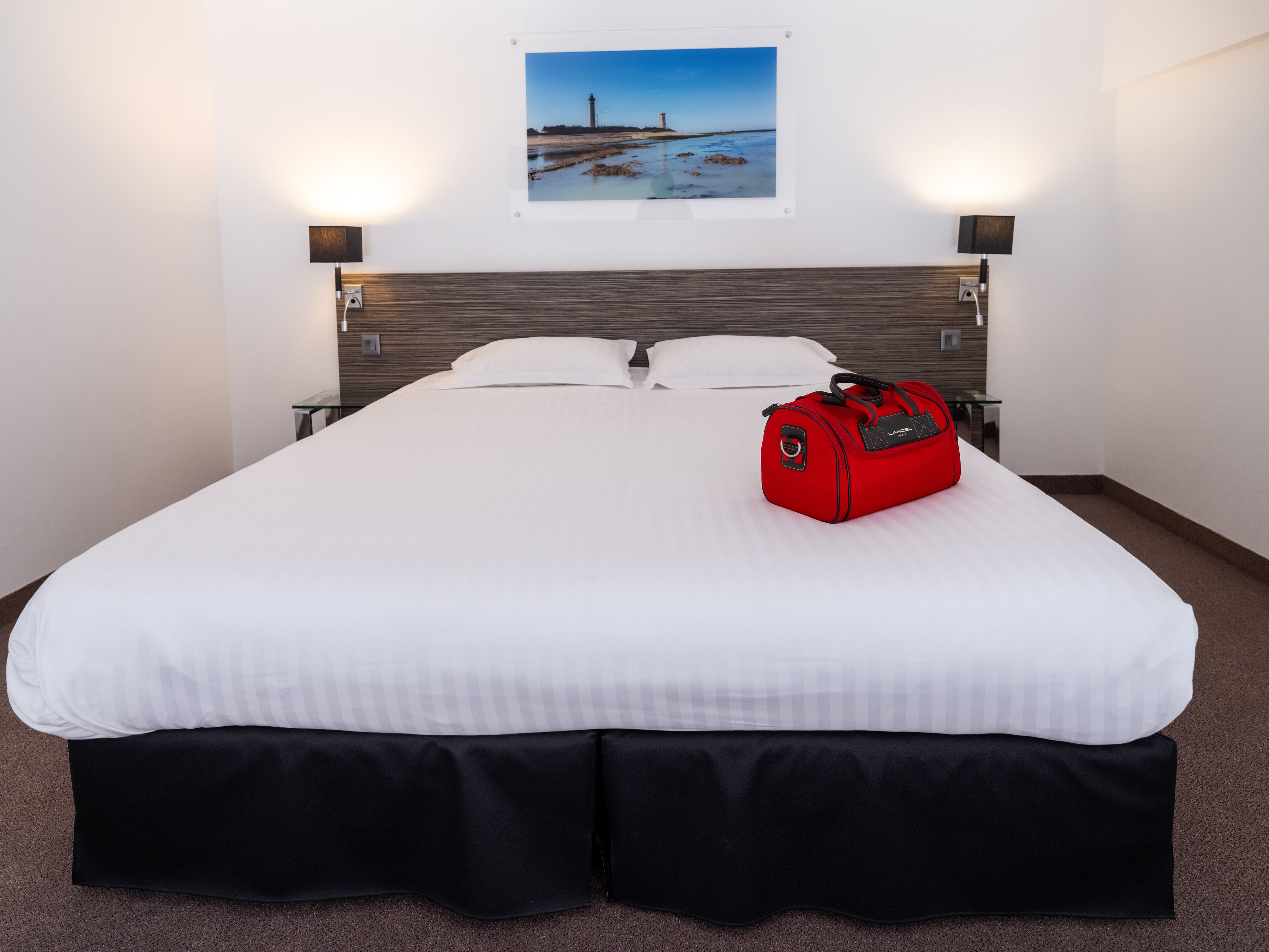 A hotel room with a large bed, a red suitcase, and a framed picture of a lighthouse on the wall.