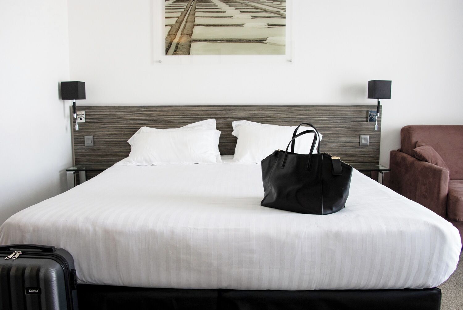 A hotel room with a bed, a black handbag on the bed, a brown chair, and a picture on the wall.