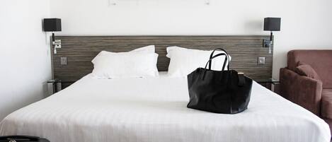 A hotel room with a bed, a black handbag on the bed, a brown chair, and a picture on the wall.