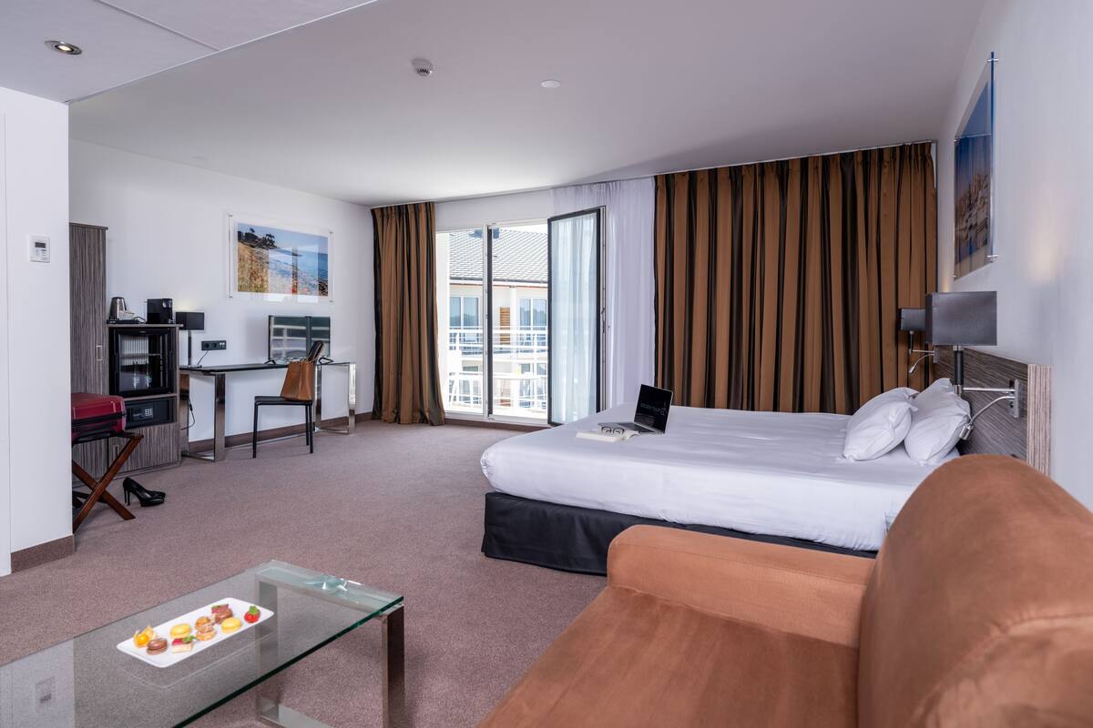 A hotel room with a bed, a desk, a chair, a glass coffee table, and a view of the city.