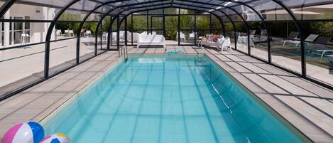 A covered swimming pool with lounge chairs and a glass enclosure.