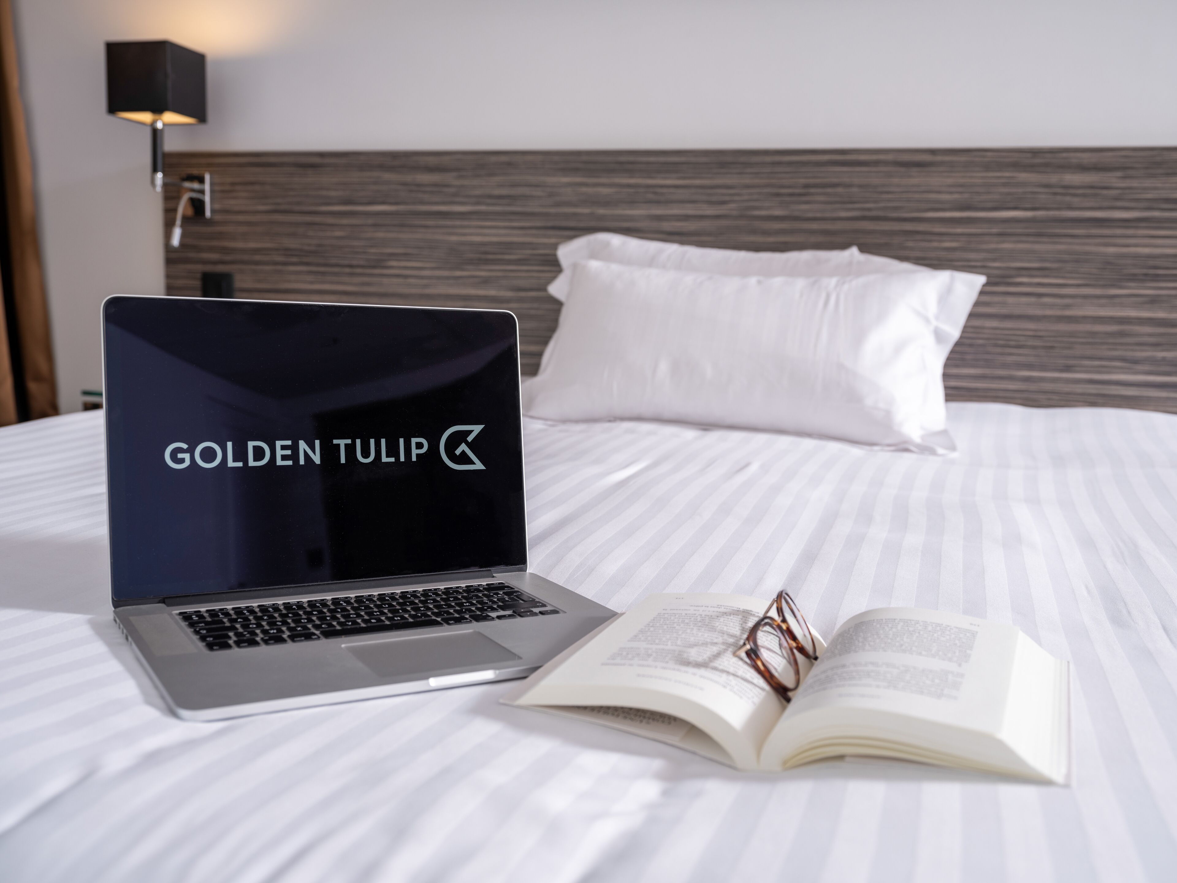Una cama con un ordenador portátil, un libro abierto y un par de gafas.