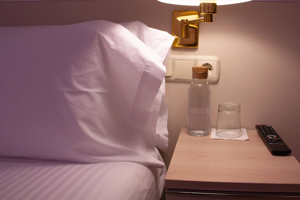 A neatly made bed with white sheets, a bedside table with a remote control, a glass bottle, and a lamp.