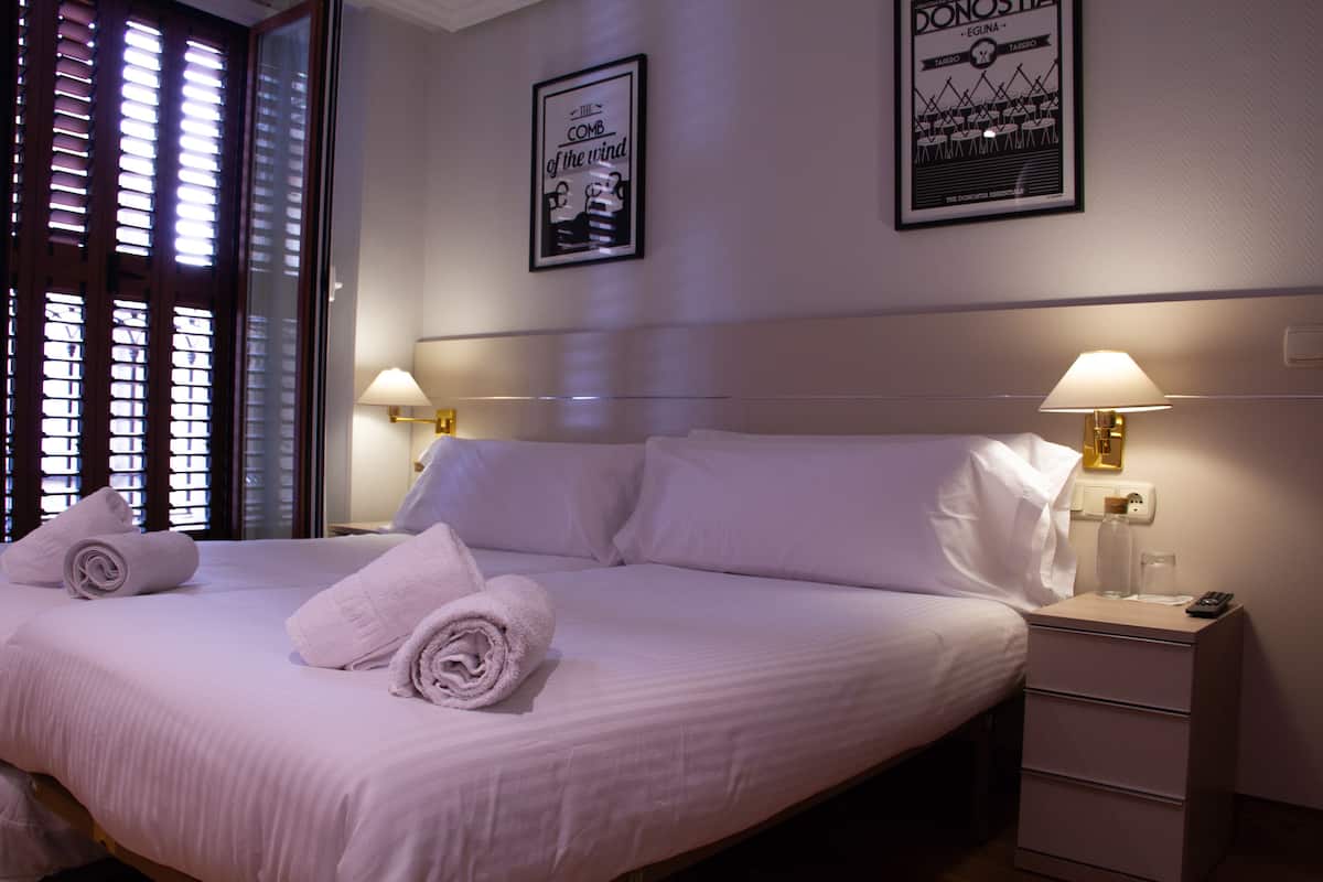 A neatly made bed with white linens and rolled towels, a bedside table with a lamp, and two framed posters on the wall.