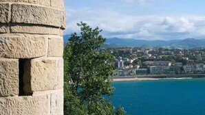 Un paisaje urbano costero con una estructura de piedra destacada a la izquierda y vistas al mar y edificios a lo lejos.
