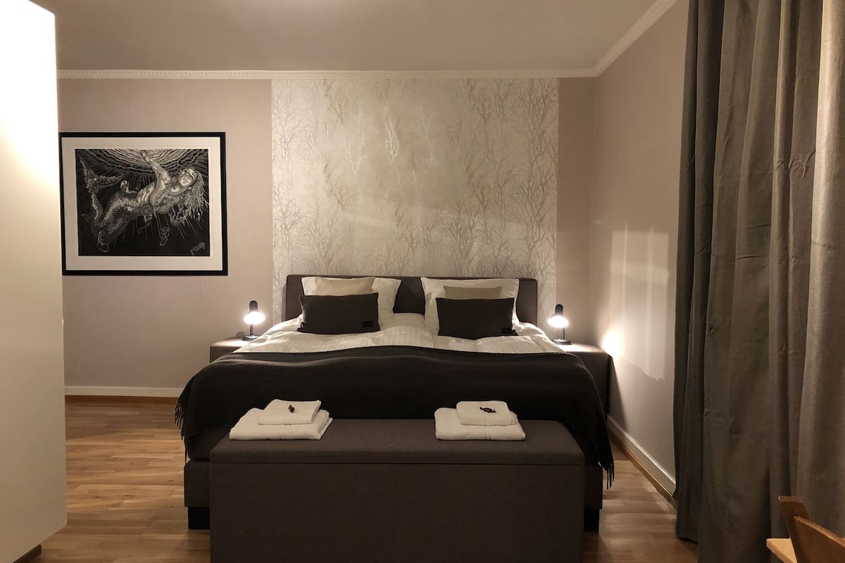 Une chambre moderne avec un grand lit, une table de chevet avec une lampe, un banc avec des serviettes et un tableau encadré au mur.