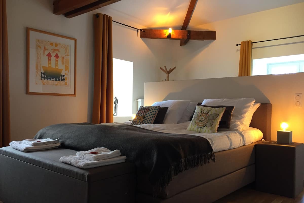 Une chambre à coucher moderne avec un grand lit, une table de chevet avec une lampe et une œuvre d’art encadrée au mur.
