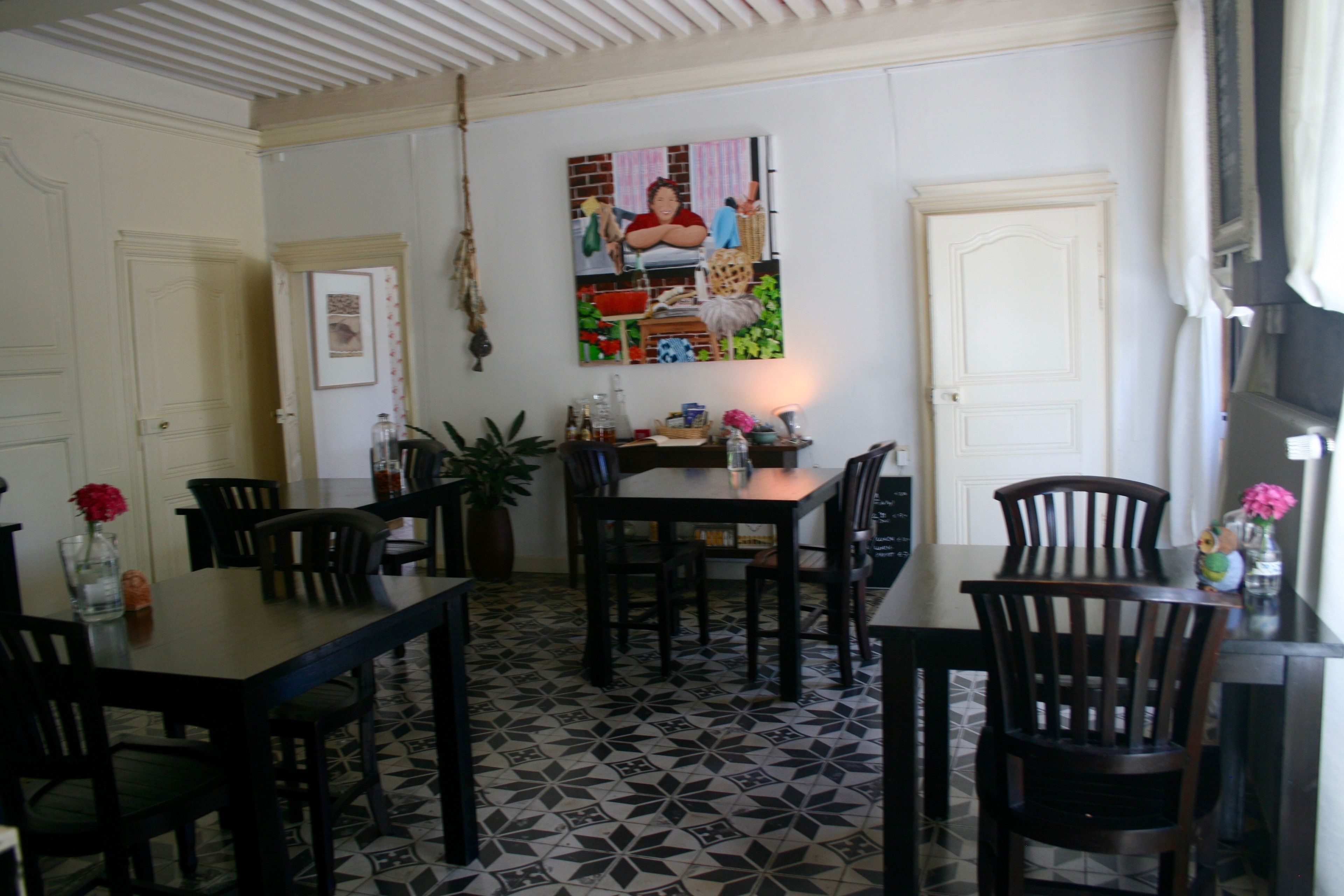 Un espace repas avec un sol à motifs, des tables et des chaises en bois, une plante suspendue et une œuvre d’art colorée accrochée au mur.