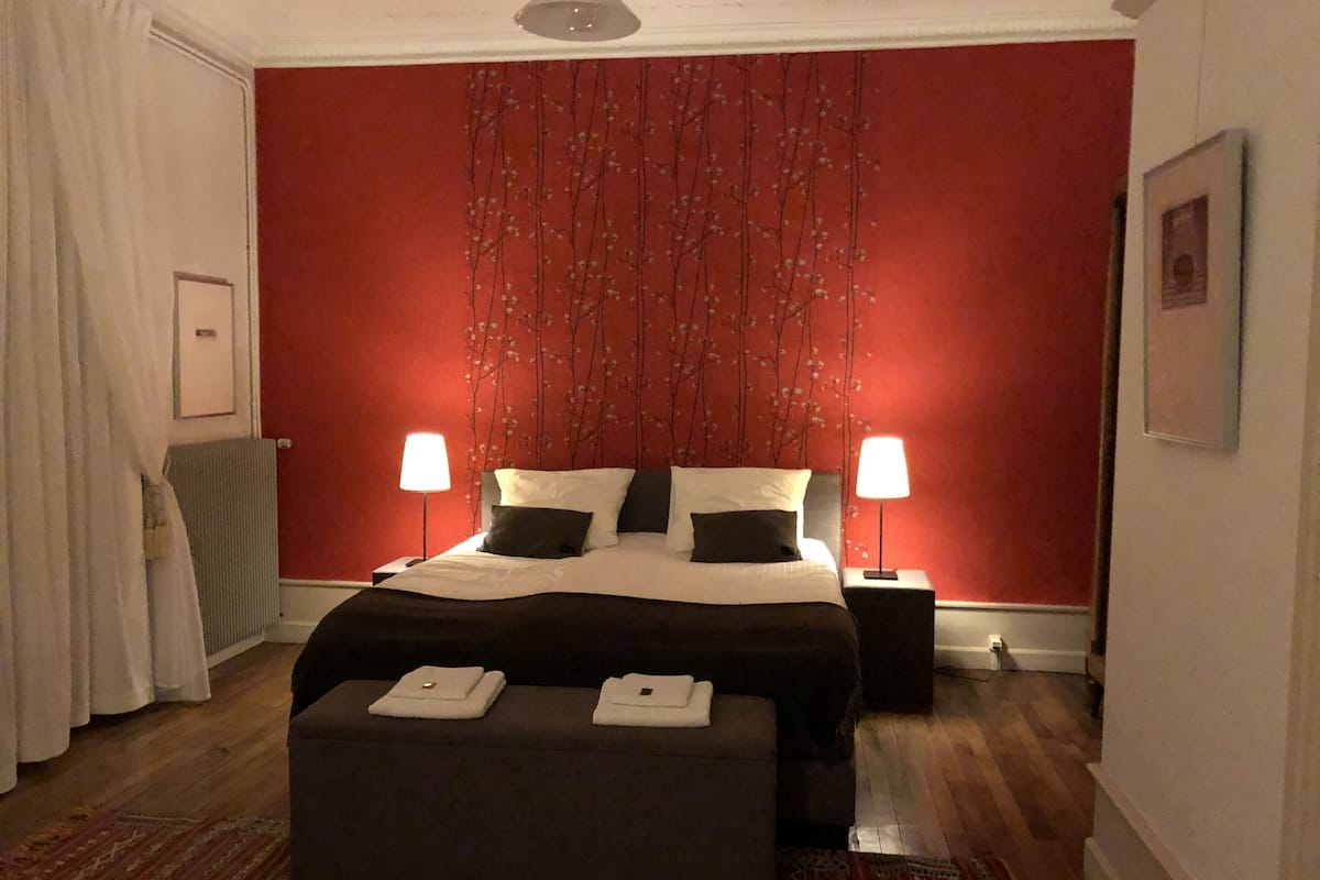 Une chambre d’hôtel avec un grand lit, deux lampes de chevet, un mur rouge orné d’un motif d’arbre, un rideau blanc et un tapis à motifs.