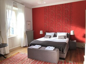 Un dormitorio con pared roja, cama con ropa de cama blanca y gris, dos lámparas de noche, un otoman gris con toallas, una alfombra estampada y una ventana con cortinas blancas.