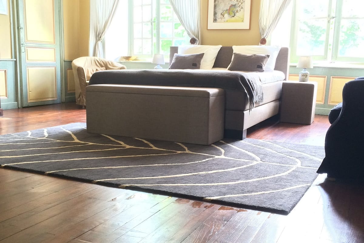 Une chambre à coucher avec un lit, une table de chevet, une chaise, un tapis et une fenêtre avec des rideaux.