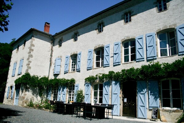 Un edificio con fachada de piedra, varias ventanas con contraventanas azules y un camino de entrada de grava.