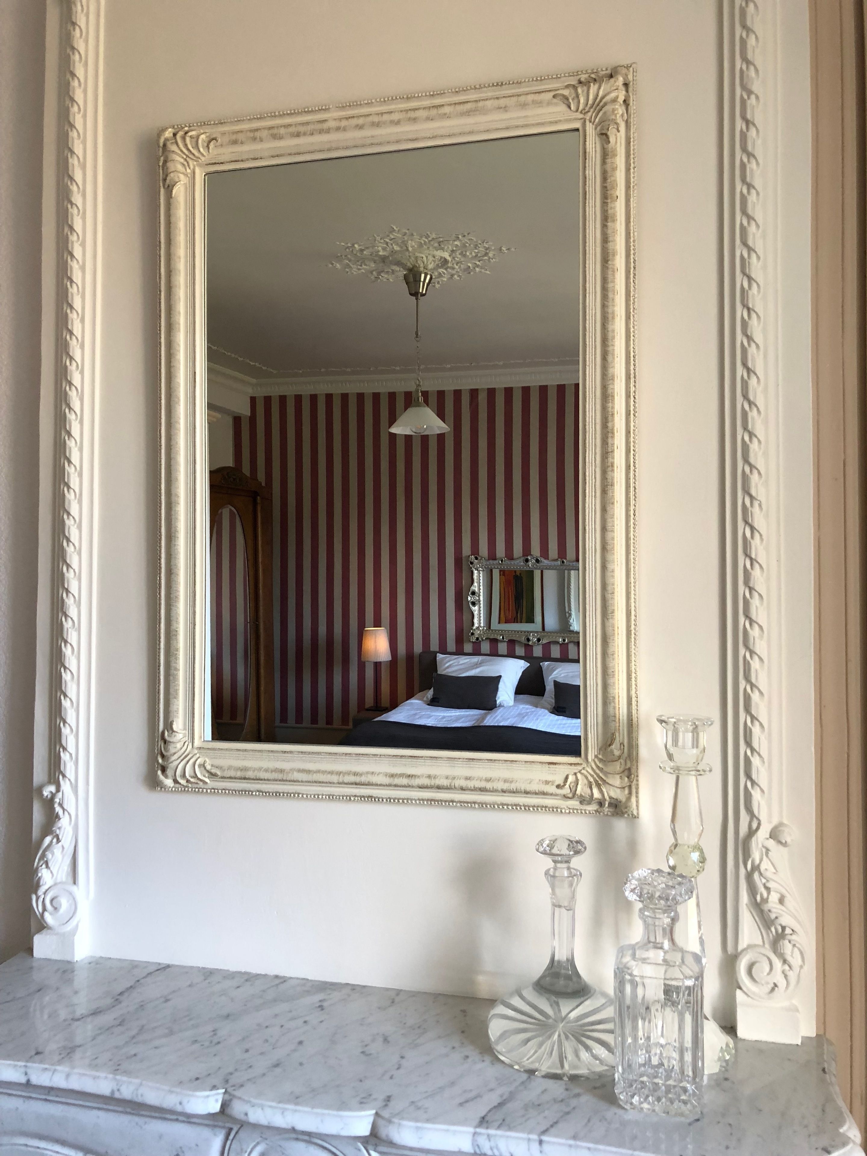 Un miroir avec un cadre décoratif, un lit recouvert d’un papier peint à rayures, une lampe et deux vases en verre posés sur une surface en marbre.