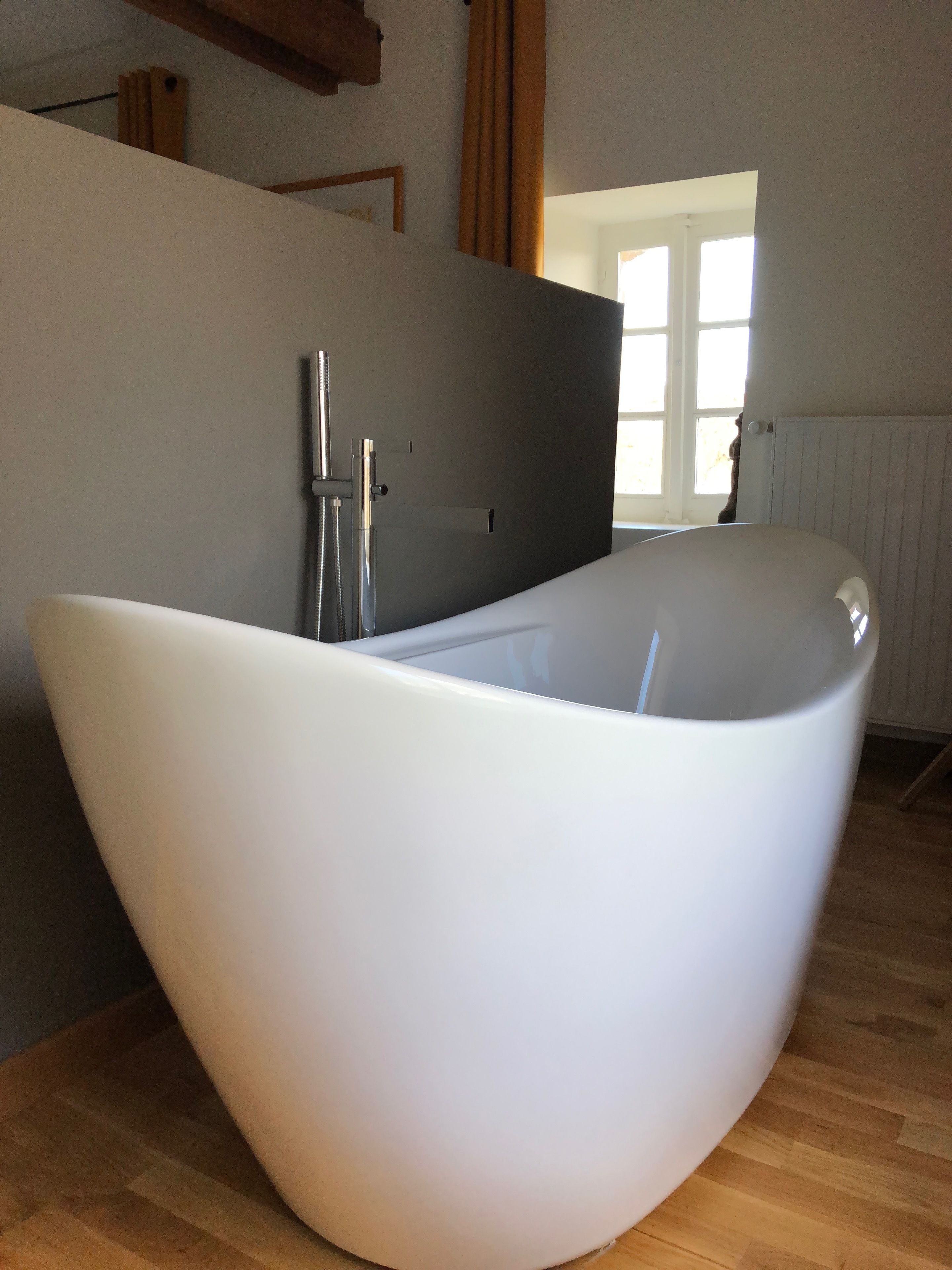 A modern bathroom with a large, white, bowl-shaped bathtub, wooden flooring, and a window providing natural light.
