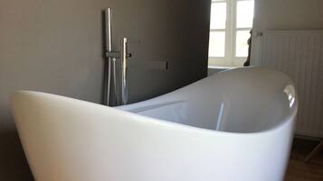 A modern bathroom with a large, white, bowl-shaped bathtub, wooden flooring, and a window providing natural light.