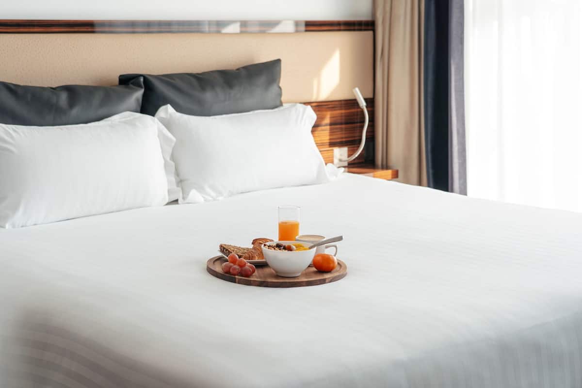 Uma cama bem arrumada com lençóis brancos, uma bandeja de madeira com itens de café da manhã e uma mesa de cabeceira com abajur.