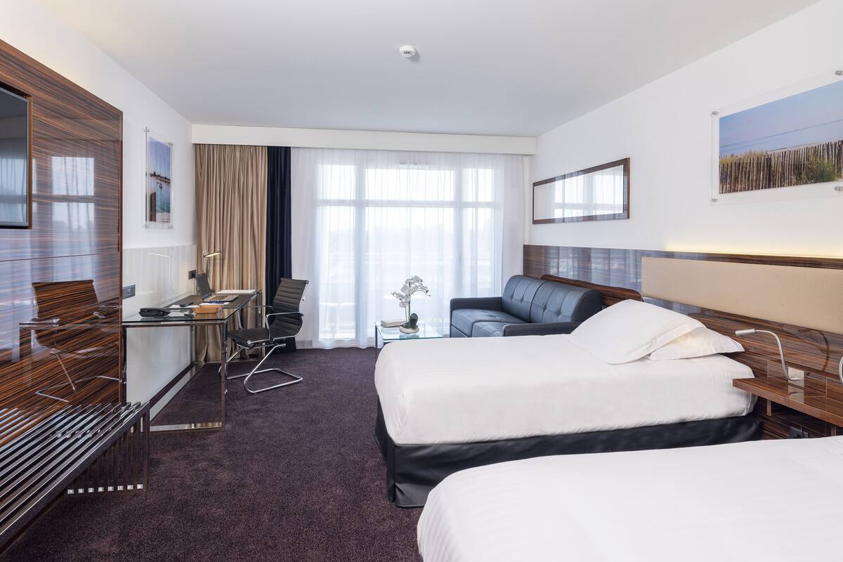 Quarto de hotel com duas camas, uma escrivaninha com cadeira, um sofá, uma televisão e uma janela grande com cortinas.
