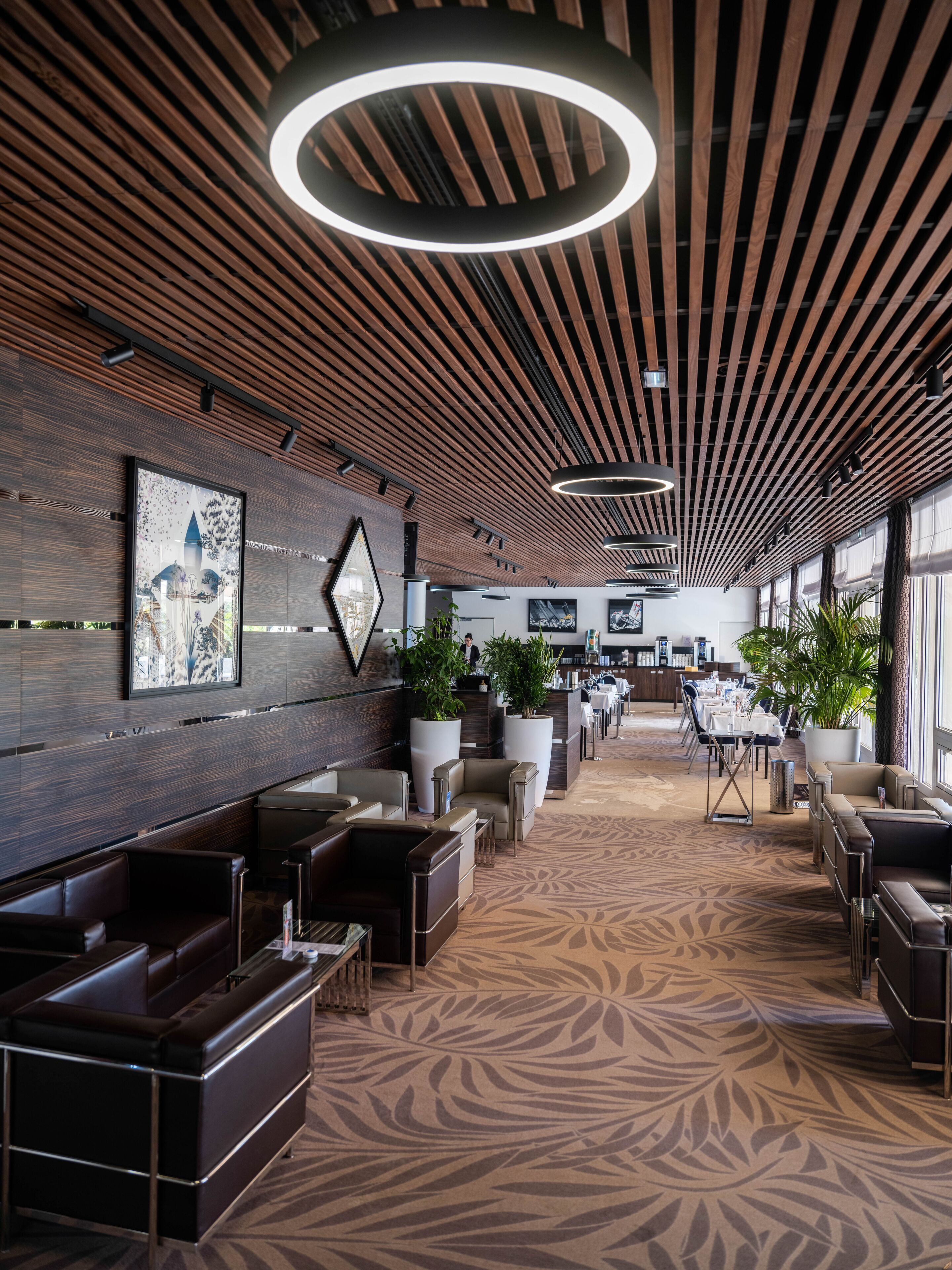 Un hall d’hôtel moderne avec un plafond en bois, de grandes plantes en pot et un tapis à motifs.