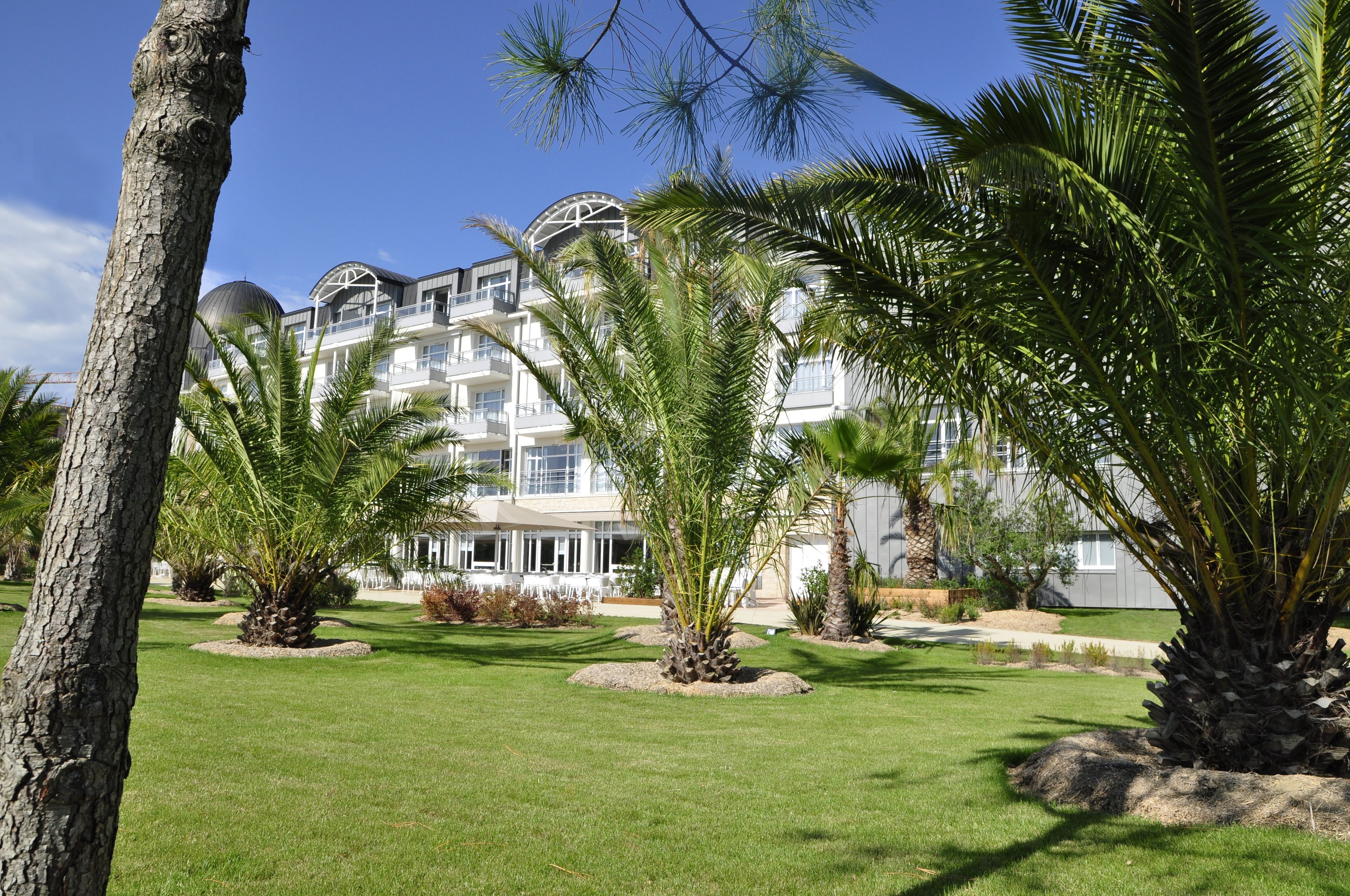 Een hotel met meerdere verdiepingen, grote ramen en een verzorgde tuin met palmbomen.
