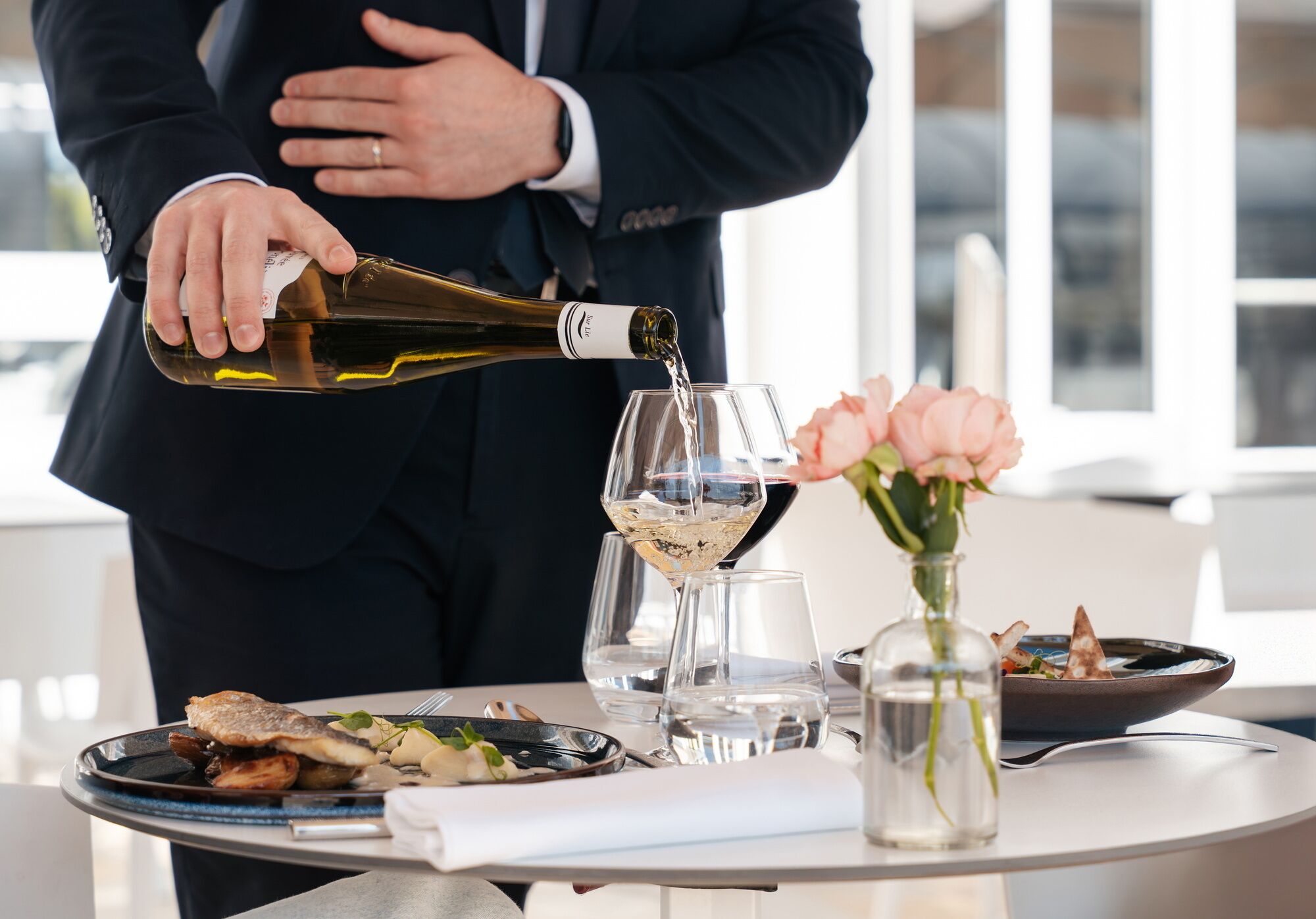 A person pouring wine into a glass, a plate of food, and a vase with flowers on a table.