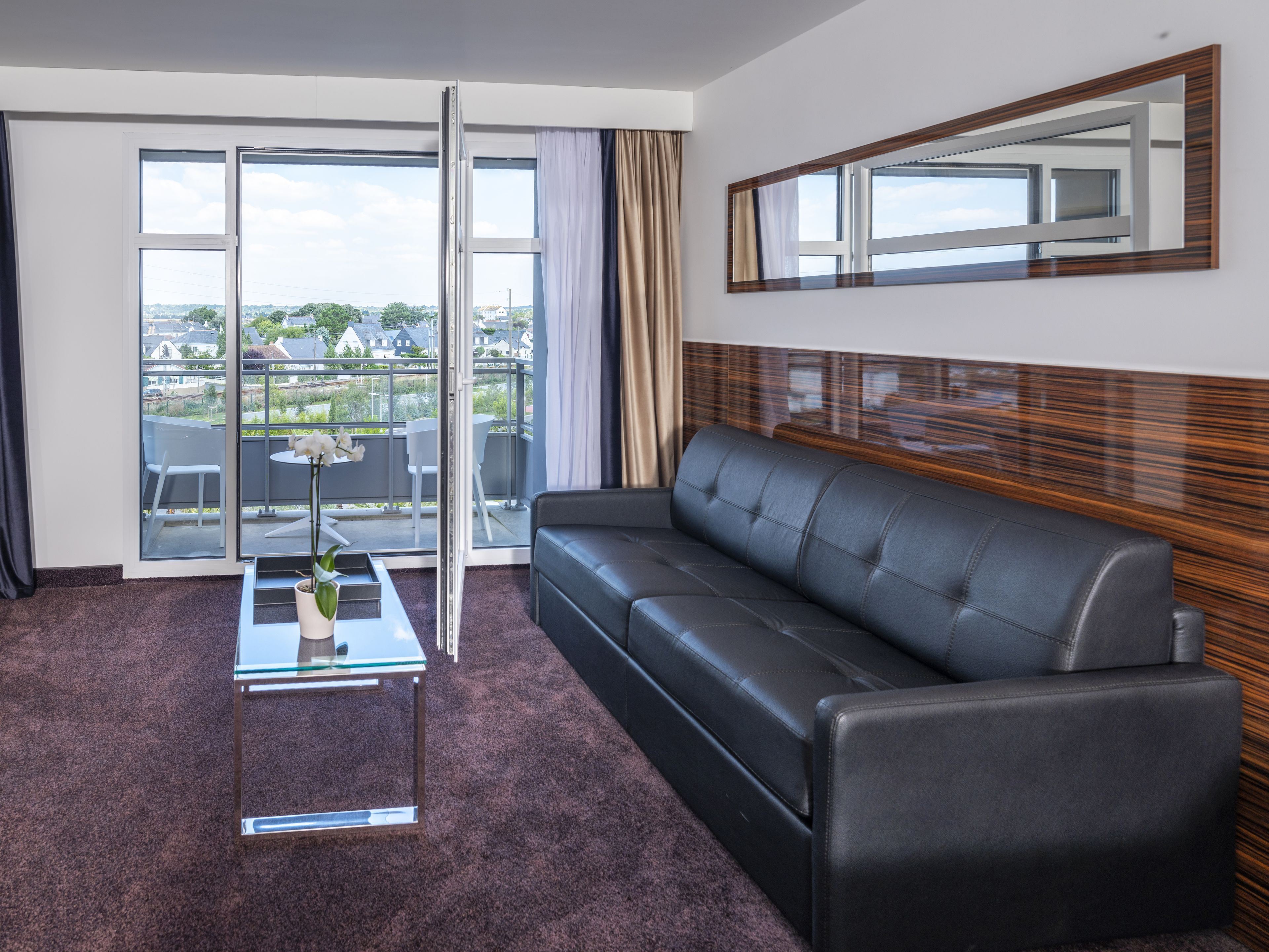 A modern living room with a black leather sofa, a glass coffee table, and a view through a glass door to the outside.