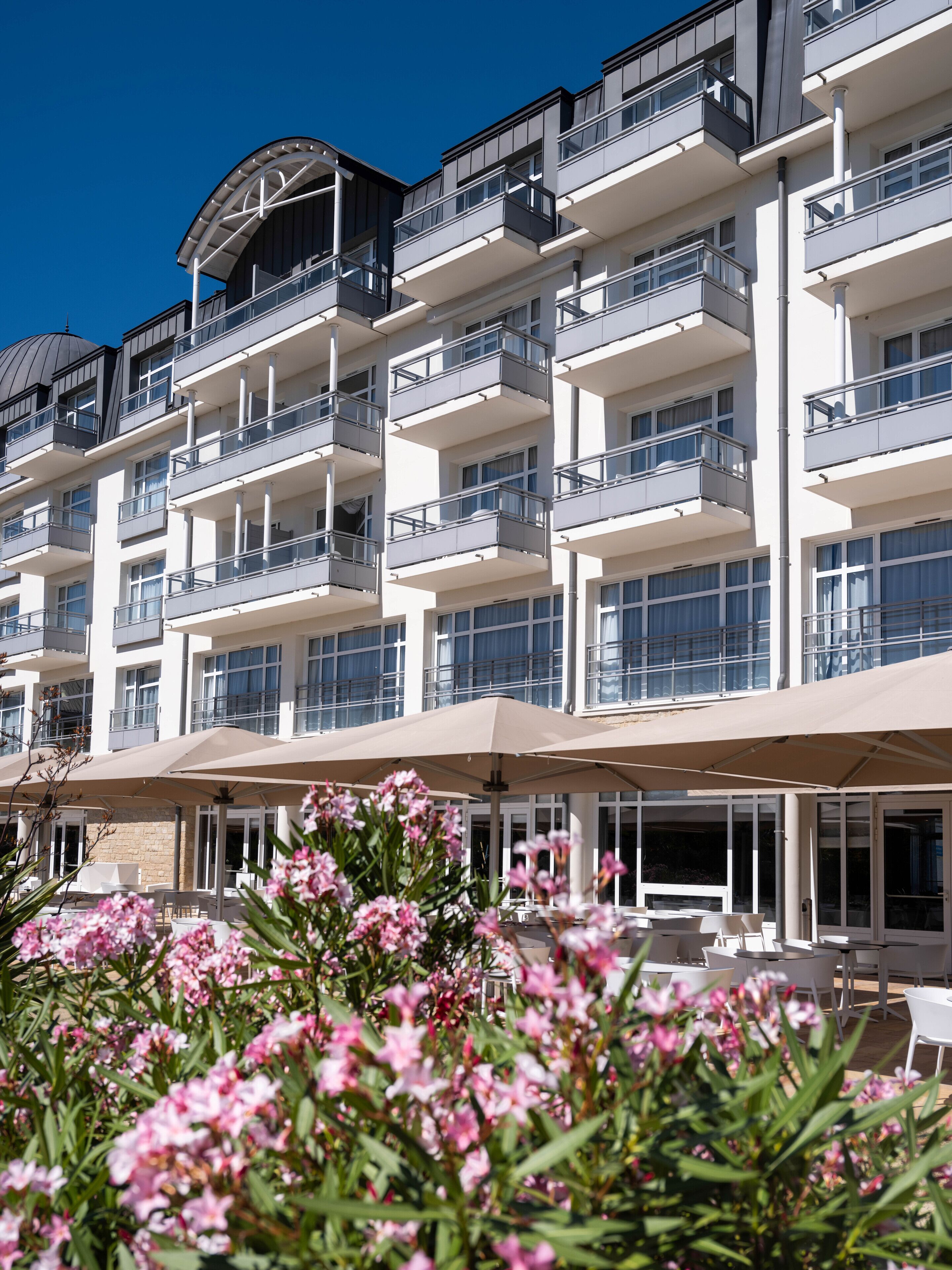 Un hotel de varios pisos con balcones, mobiliario exterior y un jardín con flores rosadas.
