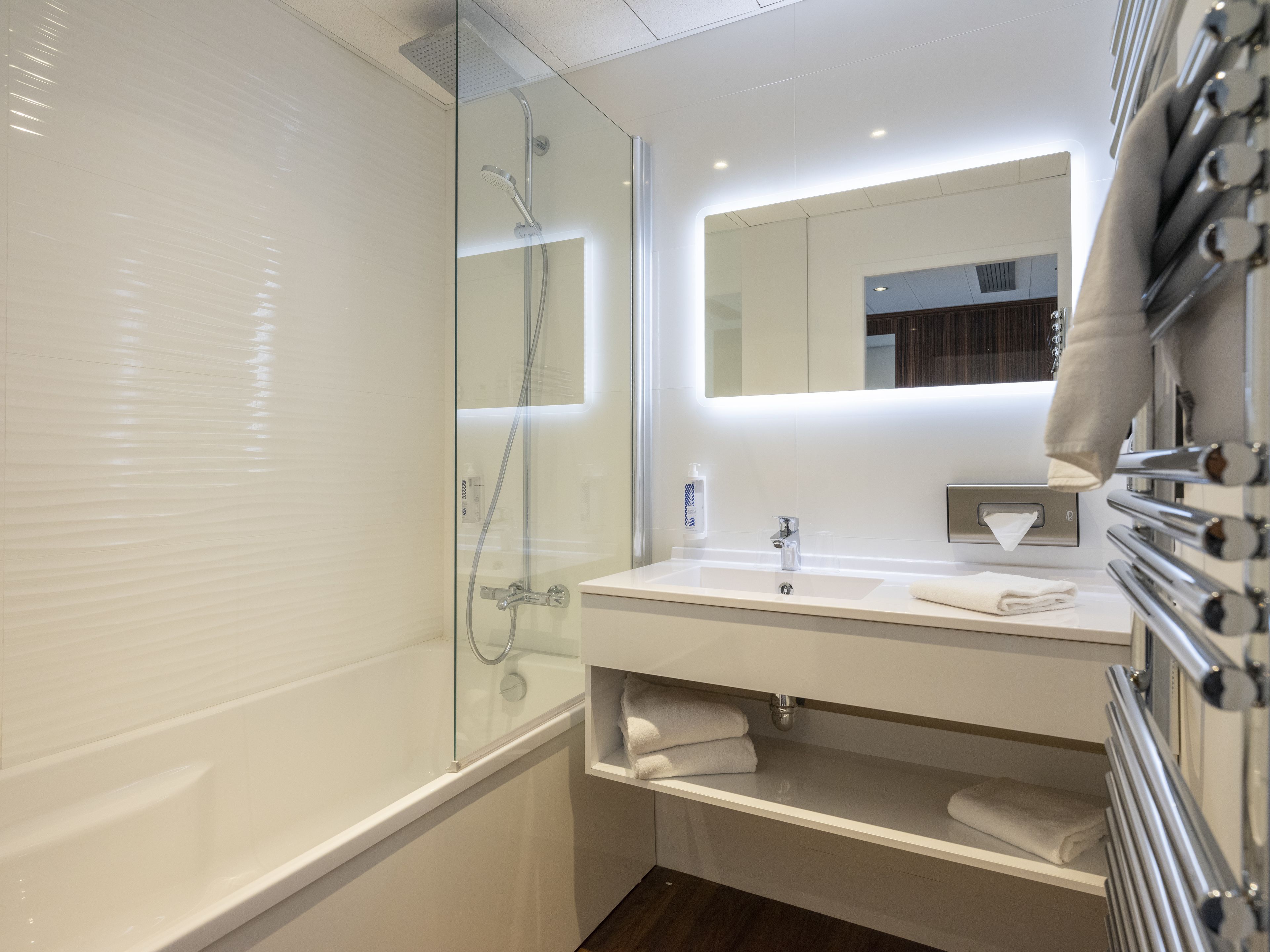 Un baño moderno con ducha de vidrio, un lavabo blanco con espejo y un perchero con toallas.