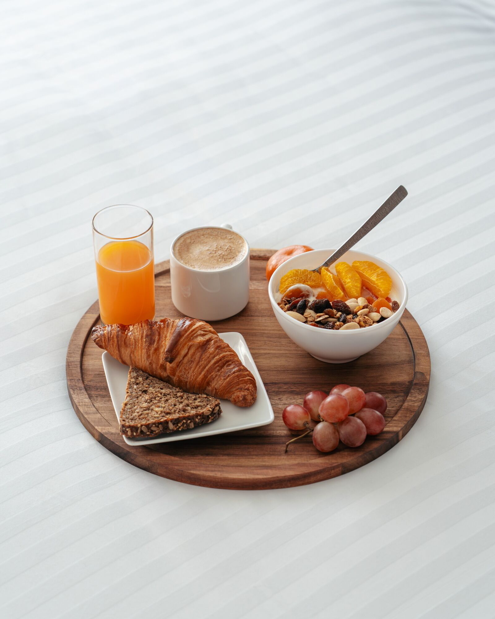 Un plateau de petit-déjeuner comprenant un croissant, une tranche de pain aux céréales, un bol de fruits et de noix, un verre de jus d’orange et une tasse de café, le tout posé sur une planche en bois.