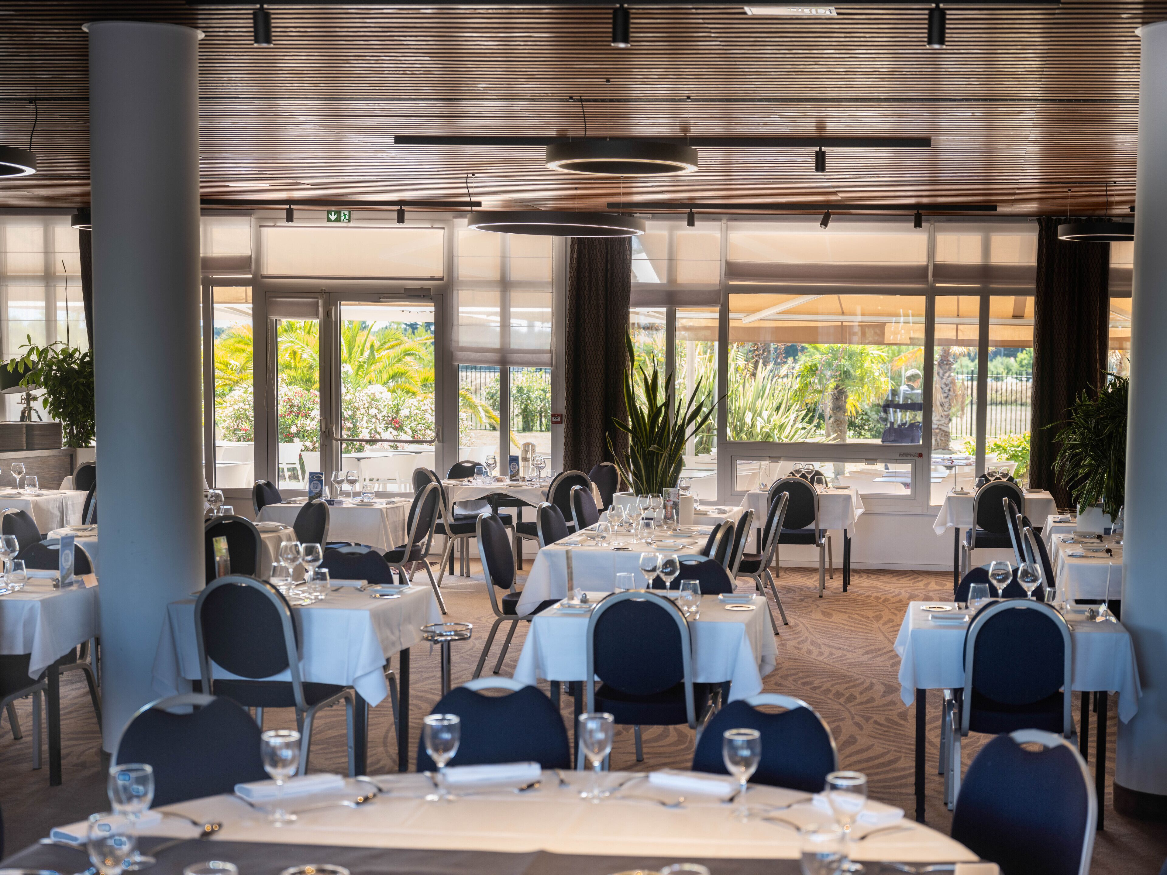 Un restaurante con varias mesas y sillas, grandes ventanales con vistas a zonas verdes y una decoración interior moderna.